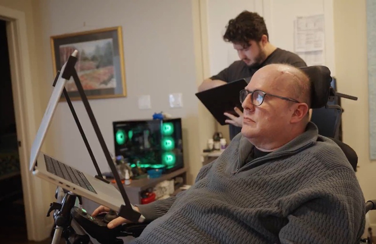 Mithilfe seines im Januar eingesetzten Neuralink-Implantats kann der stark spracheingeschränkte ALS-Patient Kenneth Shock seine Gedanken in hörbare Worte umwandeln lassen. Im Hintergrund Neuralink-Engineer Sammy Nio